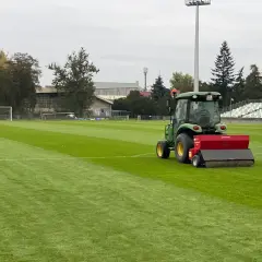 Zielony traktor z siewnikiem pracuje na boisku piłkarskim, pomagając jak zagęścić trawnik. W tle trybuny i drzewa.