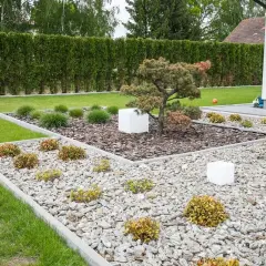 Nowoczesne klomby z kamieniami i roślinami, otoczone zielonym trawnikiem i żywopłotem. Białe kostki dodają geometrycznego charakteru.