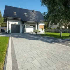 Piękny podjazd do domu z kostki brukowej, otoczony zielonym trawnikiem i drzewami.