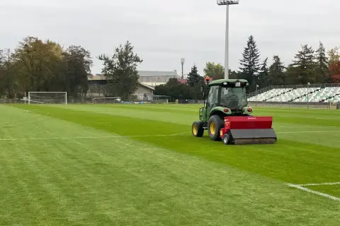 Zielony traktor z siewnikiem pracuje na boisku piłkarskim, pomagając jak zagęścić trawnik. W tle trybuny i drzewa.