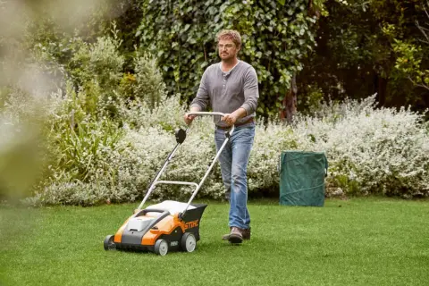 Mężczyzna z wertykulatorem Stihl dba o trawnik. W tle bujna zieleń i kwitnące krzewy.