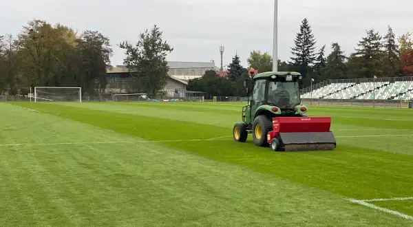 Zielony traktor z siewnikiem pracuje na boisku piłkarskim, pomagając jak zagęścić trawnik. W tle trybuny i drzewa.