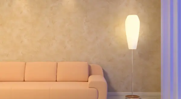 Miękka, brzoskwiniowa kanapa i elegancka lampa stojąca przy ścianie z fakturą przypominającą paca wenecka.