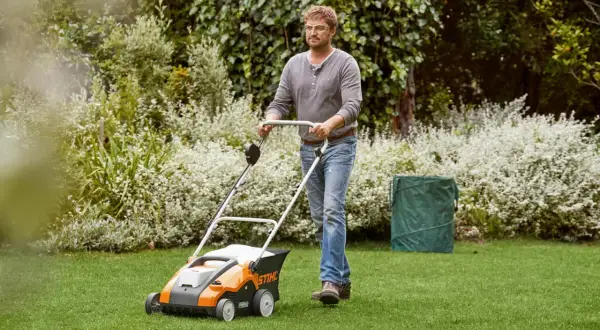 Mężczyzna z wertykulatorem Stihl dba o trawnik. W tle bujna zieleń i kwitnące krzewy.