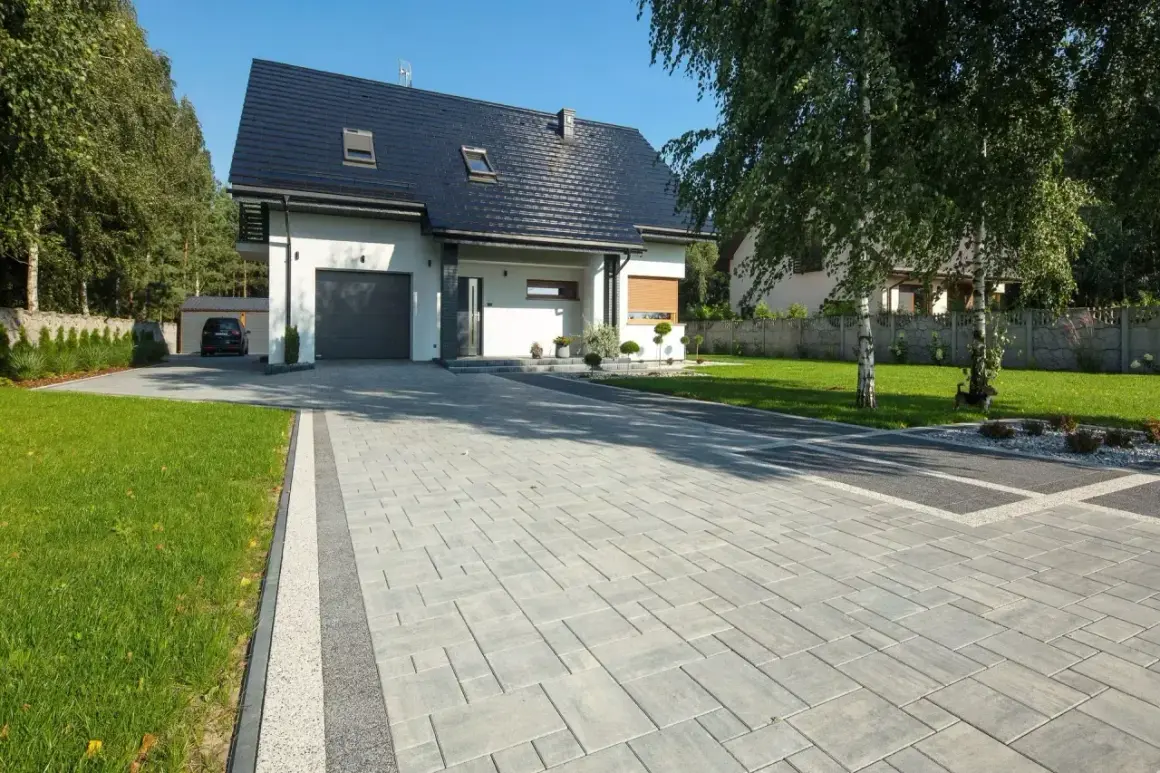 Piękny podjazd do domu z kostki brukowej, otoczony zielonym trawnikiem i drzewami.