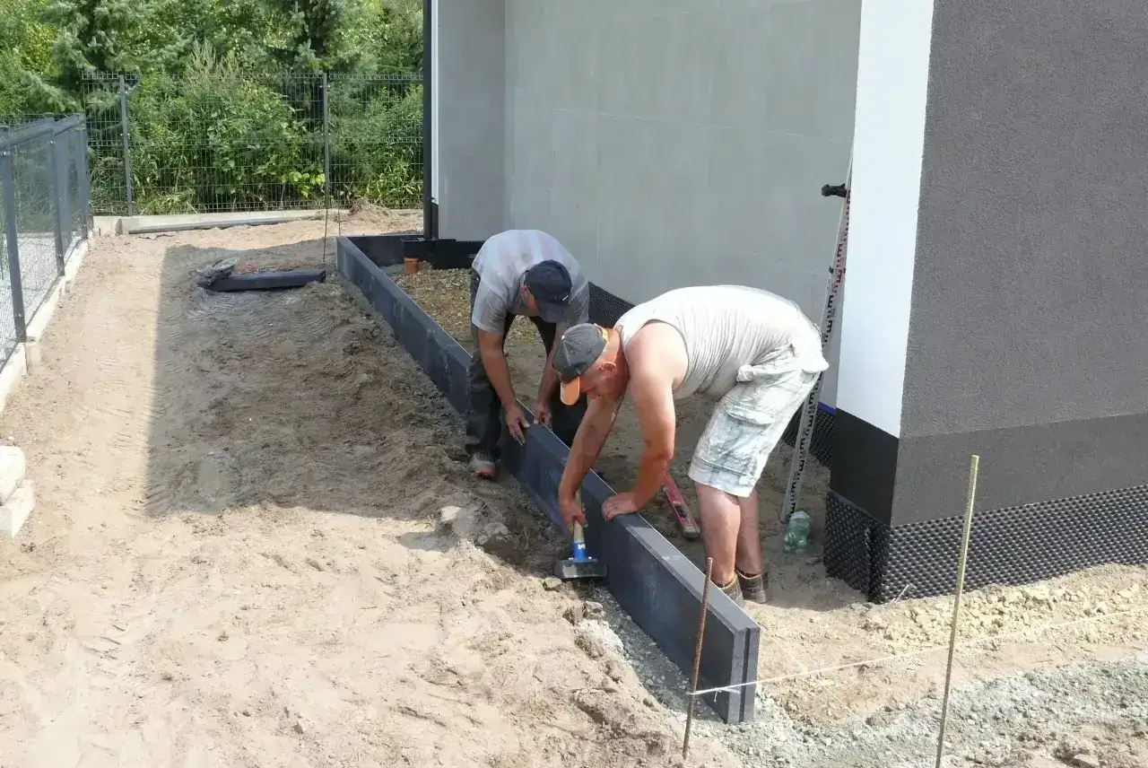 Dwóch mężczyzn układa obrzeża. W tle widać dom i ogrodzenie. Praca wymaga precyzji, jak przy dobieraniu suchy beton proporcje.