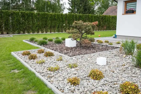 Nowoczesne klomby z kamieniami i roślinami, otoczone zielonym trawnikiem i żywopłotem. Białe kostki dodają geometrycznego charakteru.