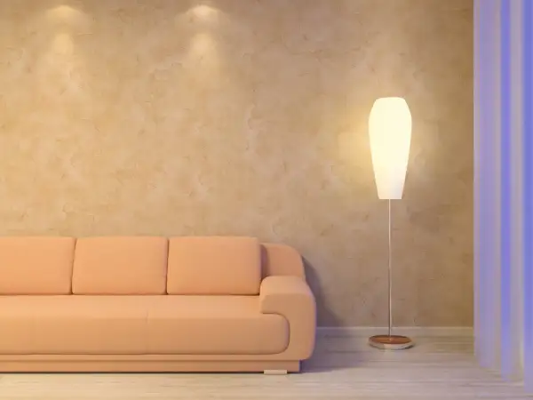 Miękka, brzoskwiniowa kanapa i elegancka lampa stojąca przy ścianie z fakturą przypominającą paca wenecka.