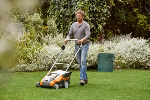 Mężczyzna z wertykulatorem Stihl dba o trawnik. W tle bujna zieleń i kwitnące krzewy.